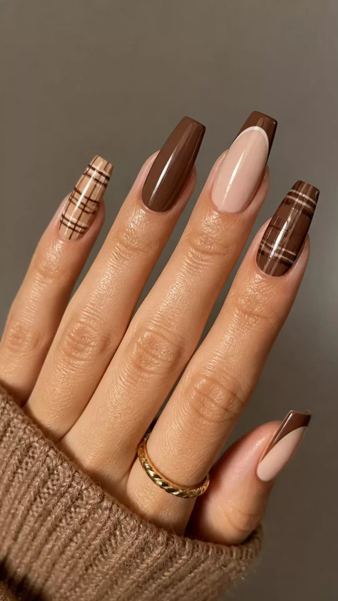 Hand with five long, manicured nails. Two nails are solid brown, two have brown and beige plaid, and one is beige with a…