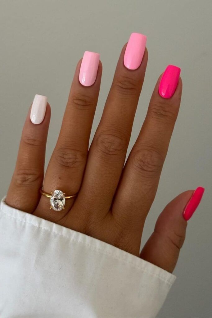 Hand with square-shaped nails in a pink ombré design. The pinky and ring finger are bright pink, the middle finger is medium…
