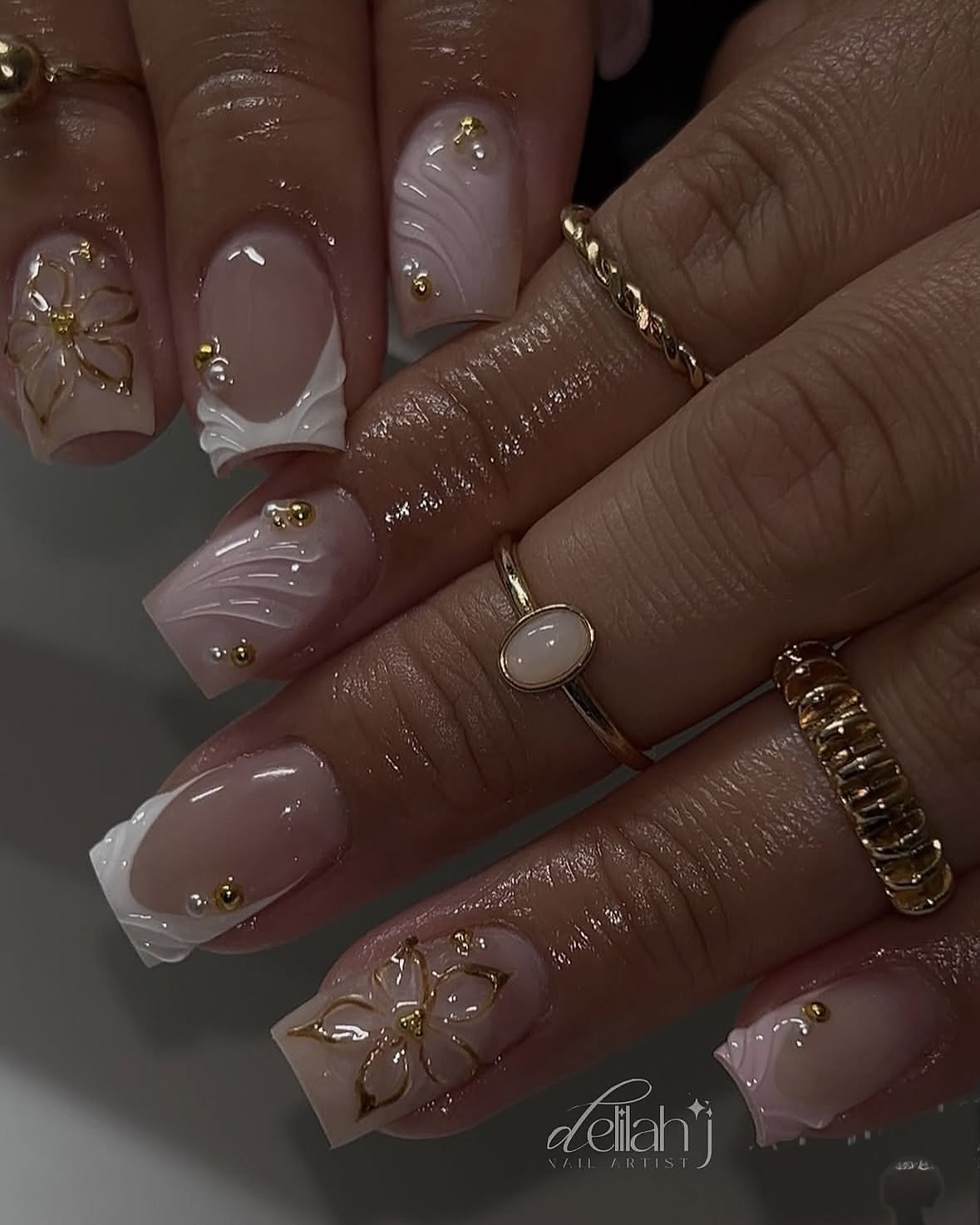 Close-up of manicured hands with short, light pink nails featuring intricate gold floral designs and wavy white accents.