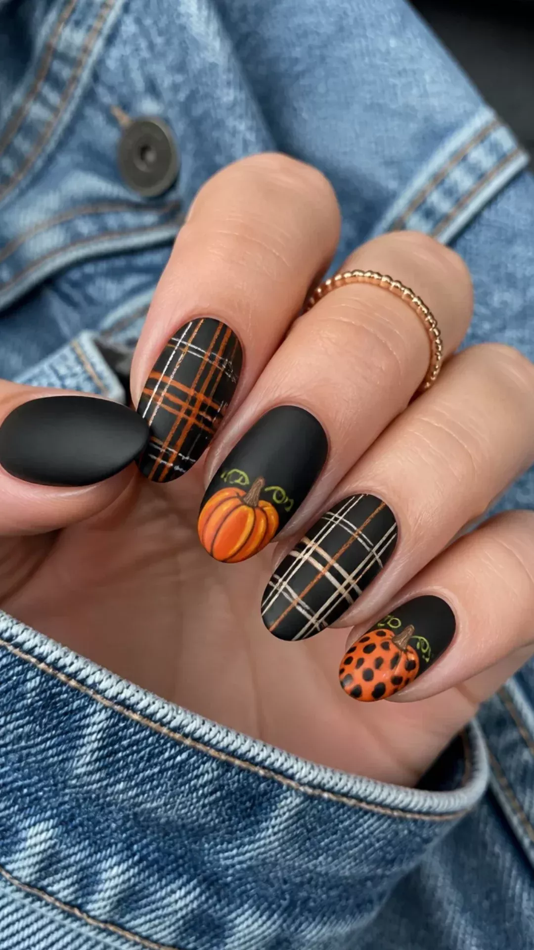 Hand with five oval-shaped nails, three with black and orange plaid, two with black matte and pumpkin art