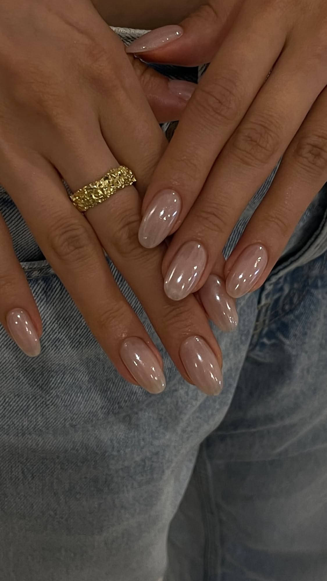 Close-up of hands with oval, iridescent nude nails, resting on light blue denim fabric.
