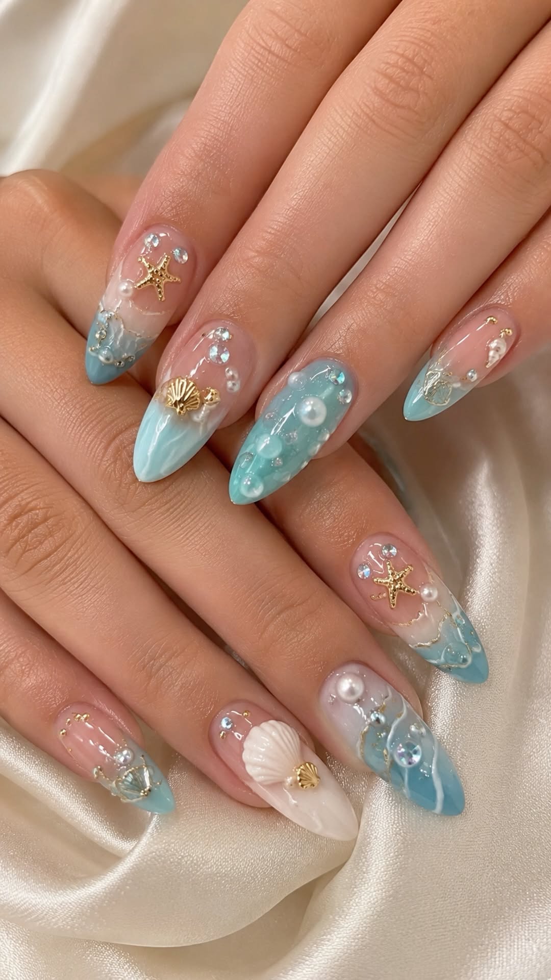 Pointed nails with blue and white ombre base and various 3D shell, starfish, pearl, and gem embellishments.