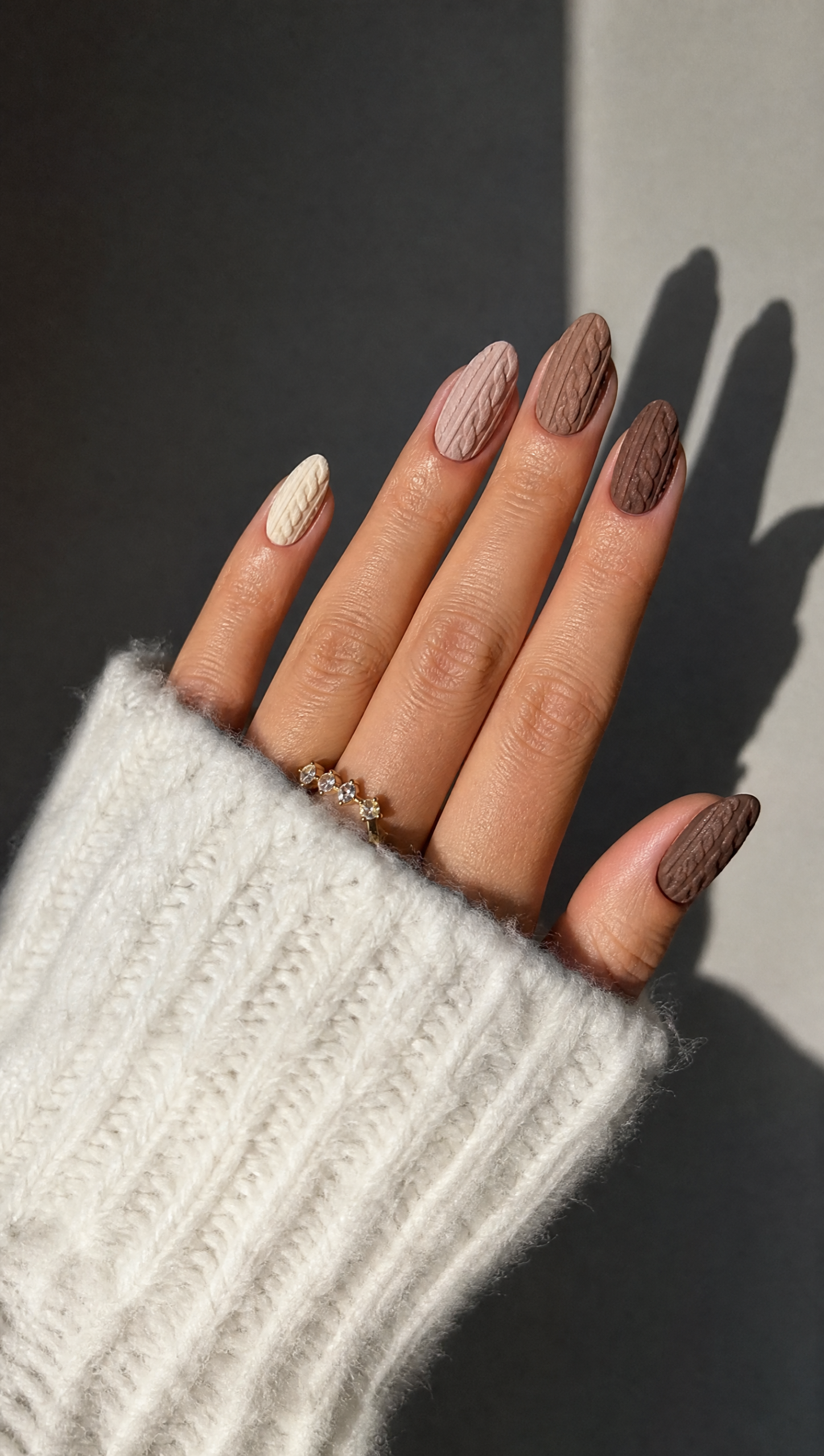 Hand with five nails, each painted with a distinct brown shade and a raised cable-knit texture, resting on a white sweater.