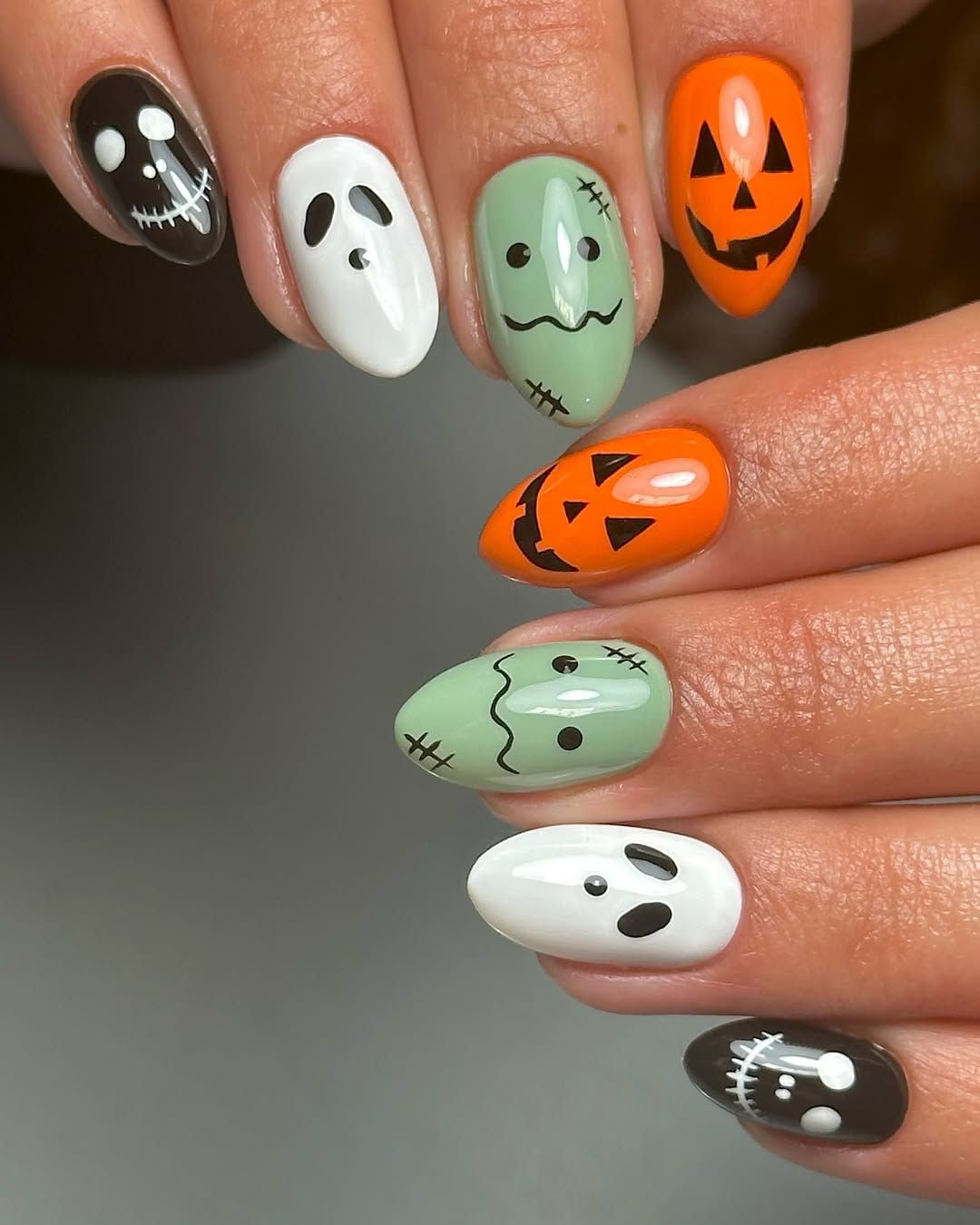Almond-shaped nails decorated with Halloween characters, including ghosts, Frankenstein's monster, and jack-o'-lanterns.