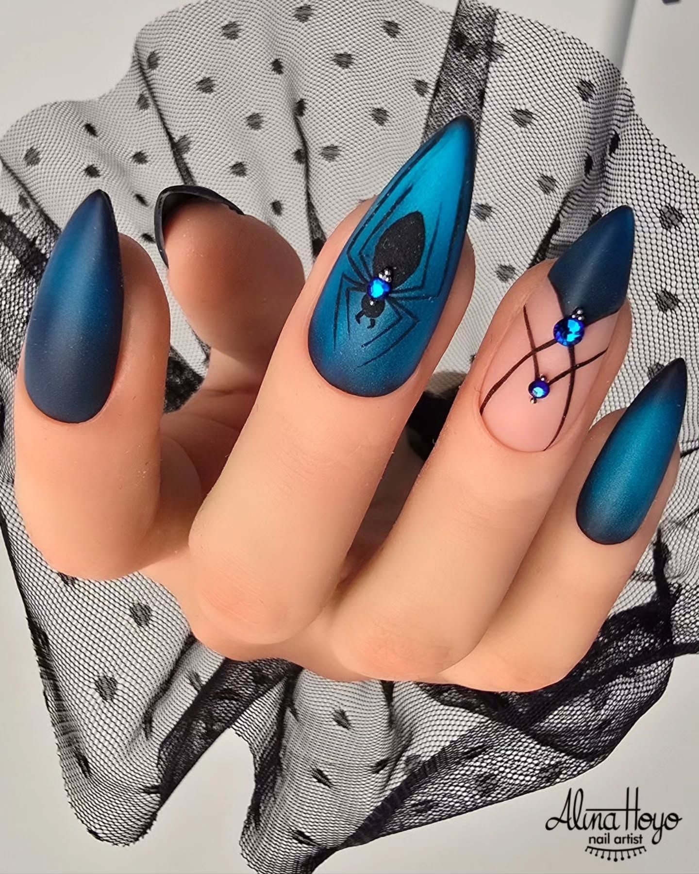 Close-up of pointed nails with blue matte and ombre polish, one nail featuring a black spider design with blue gems, another…