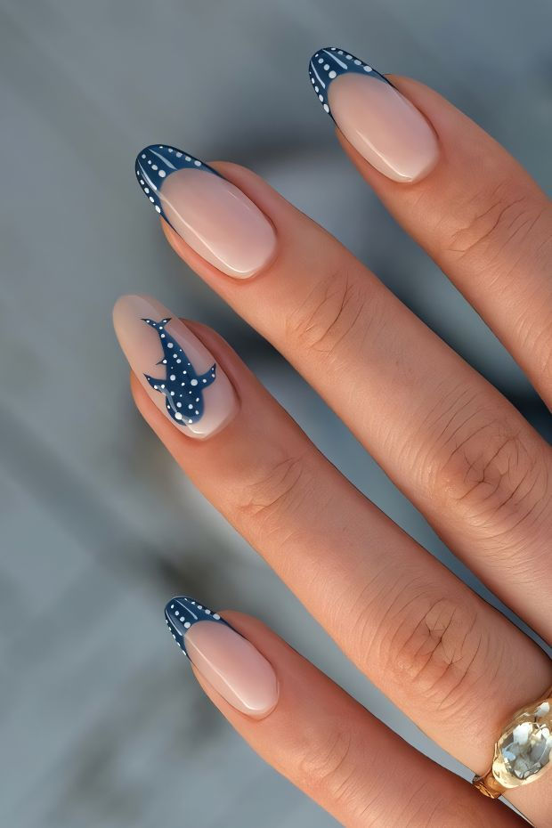 Close-up of a hand with almond-shaped nails, featuring a dark blue French tip with white dots and a whale design on one nail.