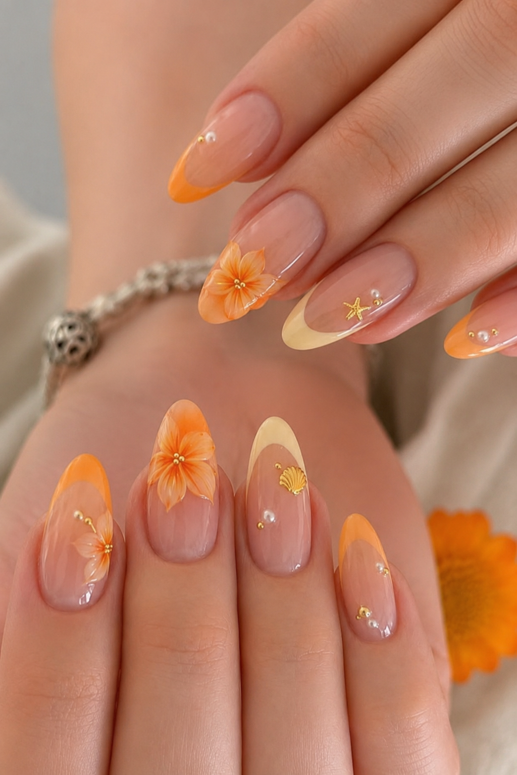 Close-up of a hand with almond-shaped nails featuring orange floral designs, seashell and starfish accents, and pearls.