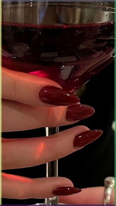 Hand with deep red almond-shaped nails holding a wine glass with red liquid.