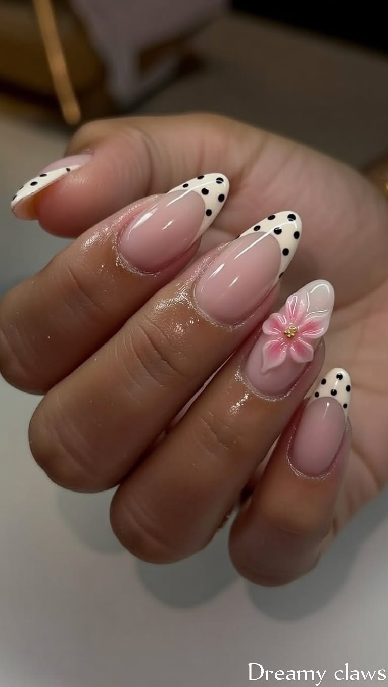 Almond-shaped nails with a sheer pink base, white French tips with black polka dots, and one nail featuring a pink 3D flower…