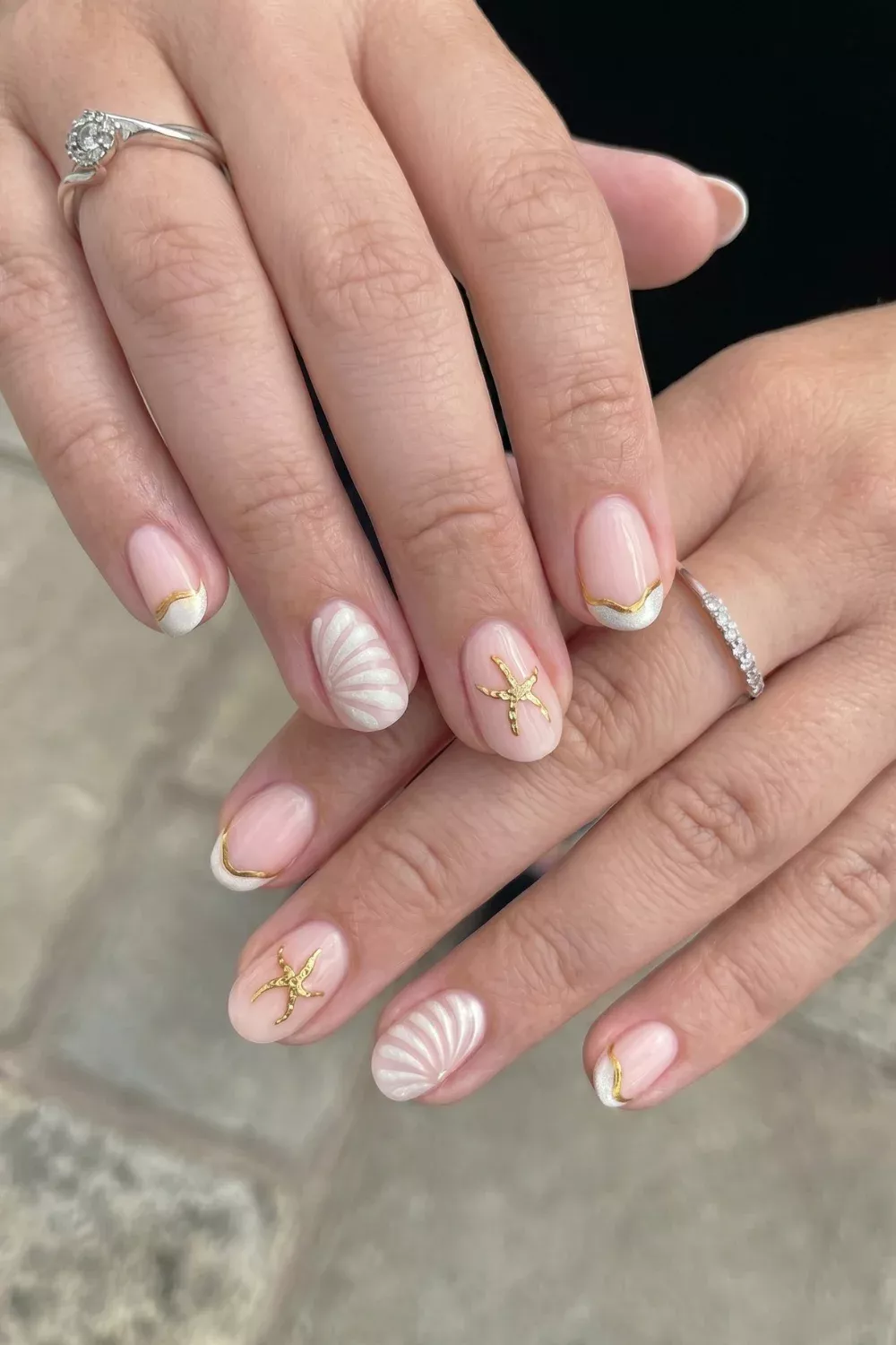 Manicured hands with short, light pink nails featuring golden starfish and white seashell designs, and curved white tips…