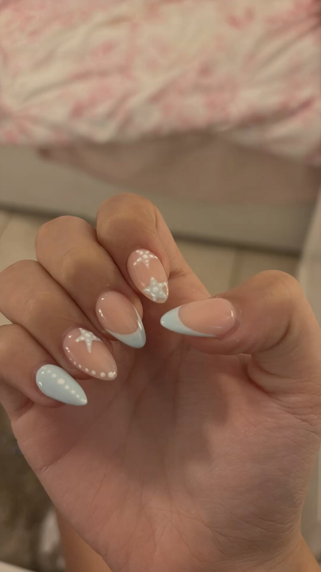 Close-up of a hand with almond-shaped nails featuring pale blue French tips and white star and dot designs.