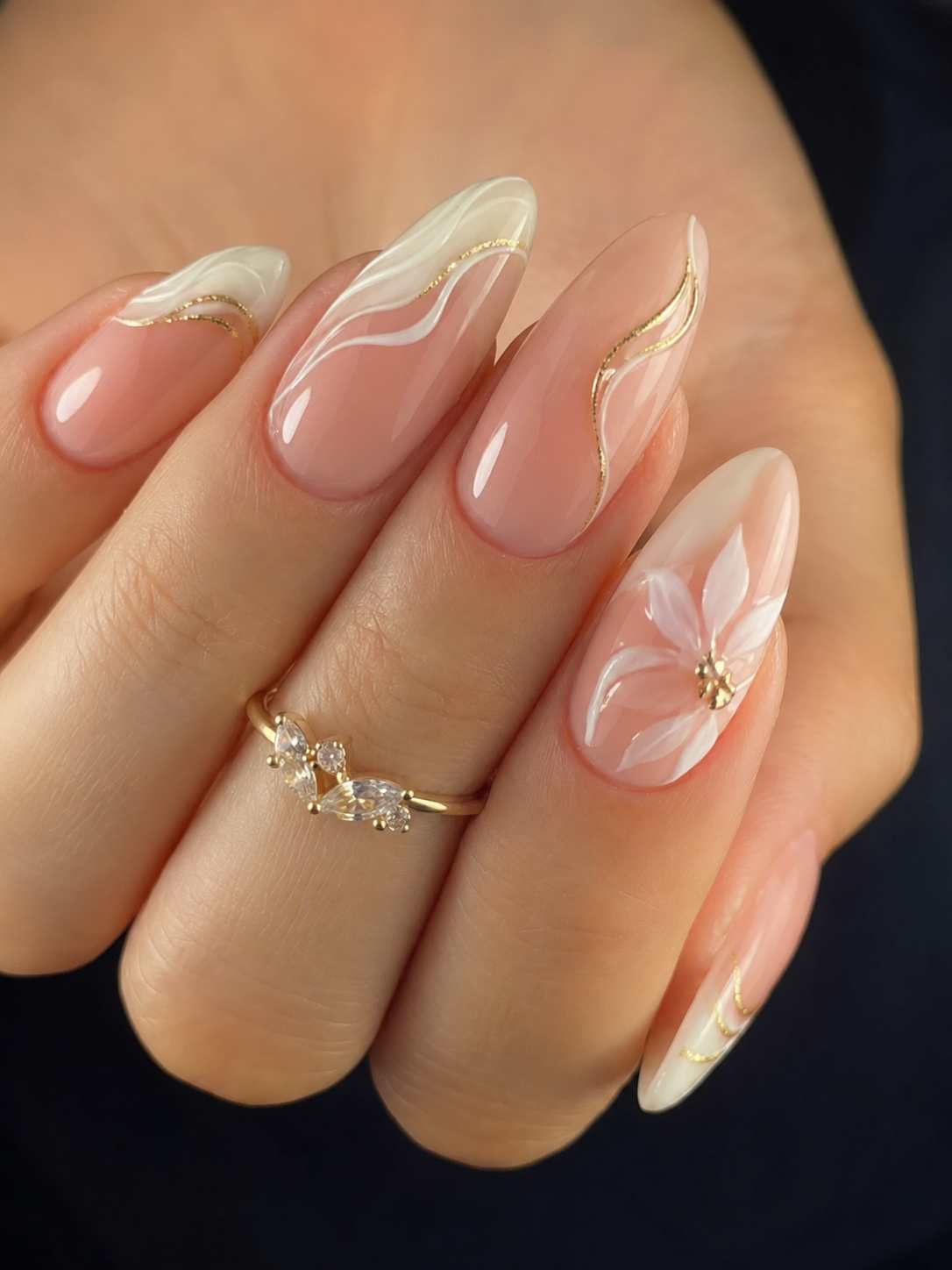Close-up of a hand with almond-shaped nails featuring a French manicure style with gold line accents and a white floral…