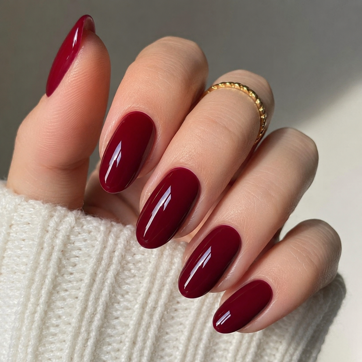 Hand with five nails polished in glossy deep red, resting against a cream knit sweater, wearing a gold ring