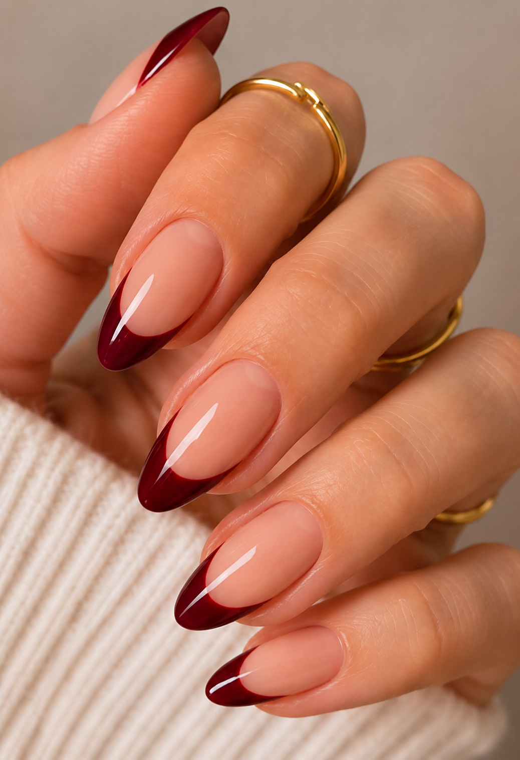 Hand with long almond-shaped nails featuring burgundy French tips over a nude base, wearing gold rings.
