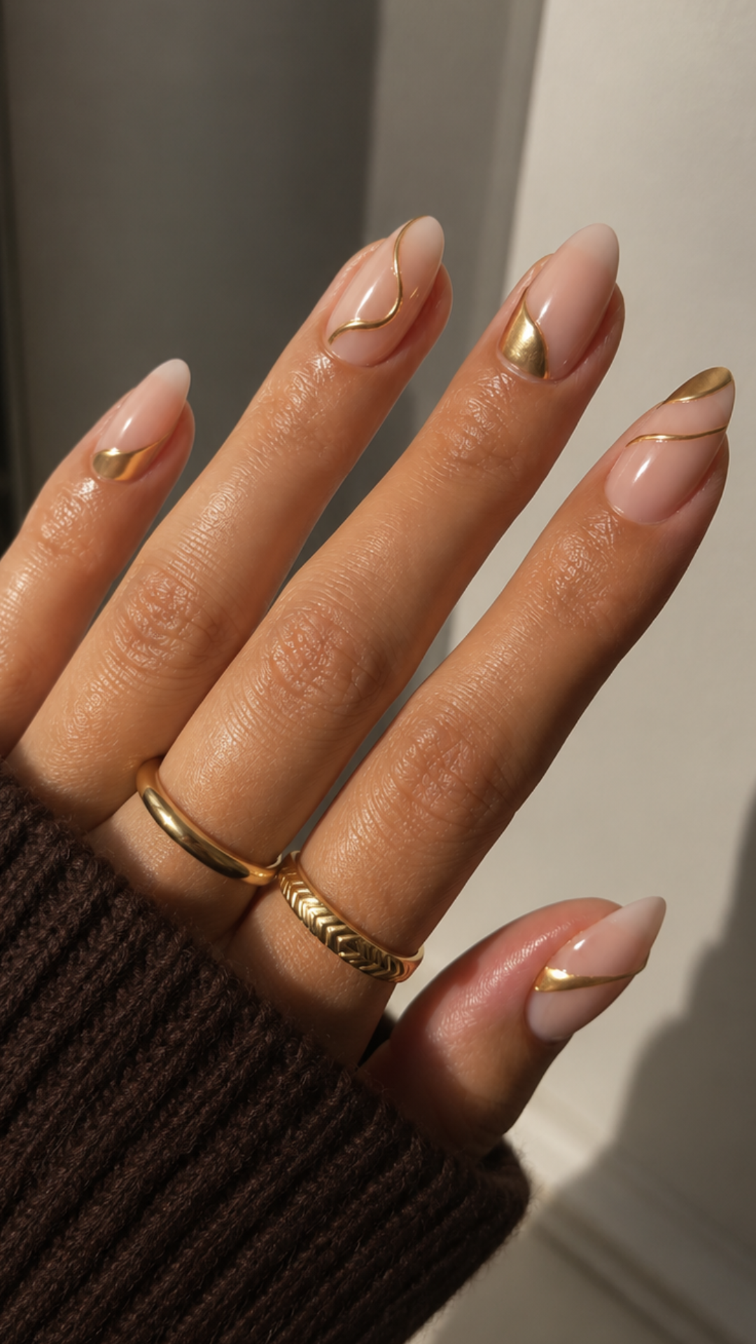 Hand with almond-shaped nails featuring minimalist gold line designs, wearing two gold rings.