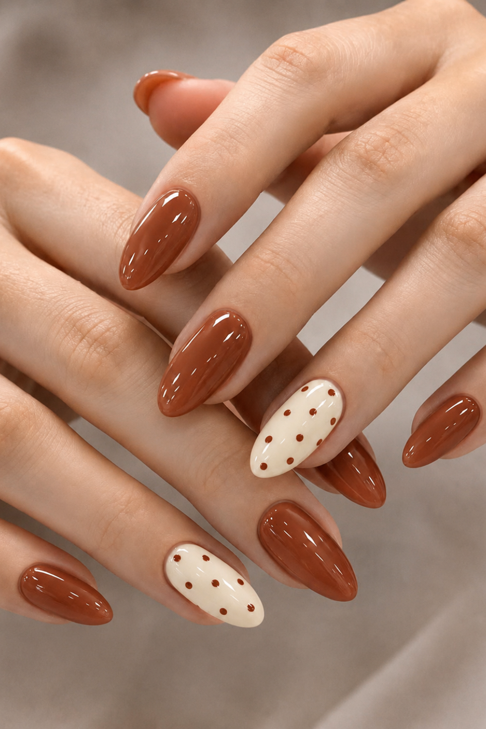 Close up of a person's hands with almond-shaped nails. Most nails are a glossy terracotta shade, and two accent nails are…