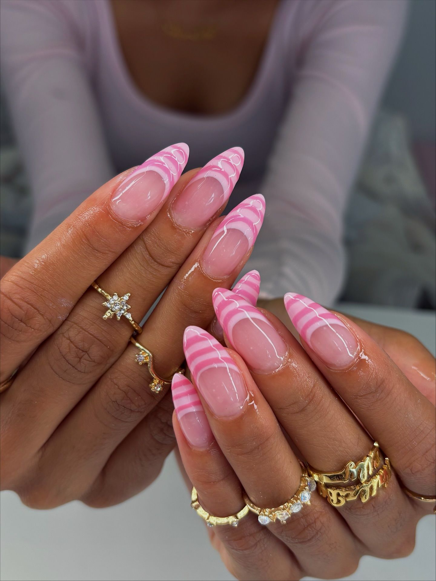 Pointed acrylic nails with pink and white stripes on the tips, worn by a person with several gold rings.