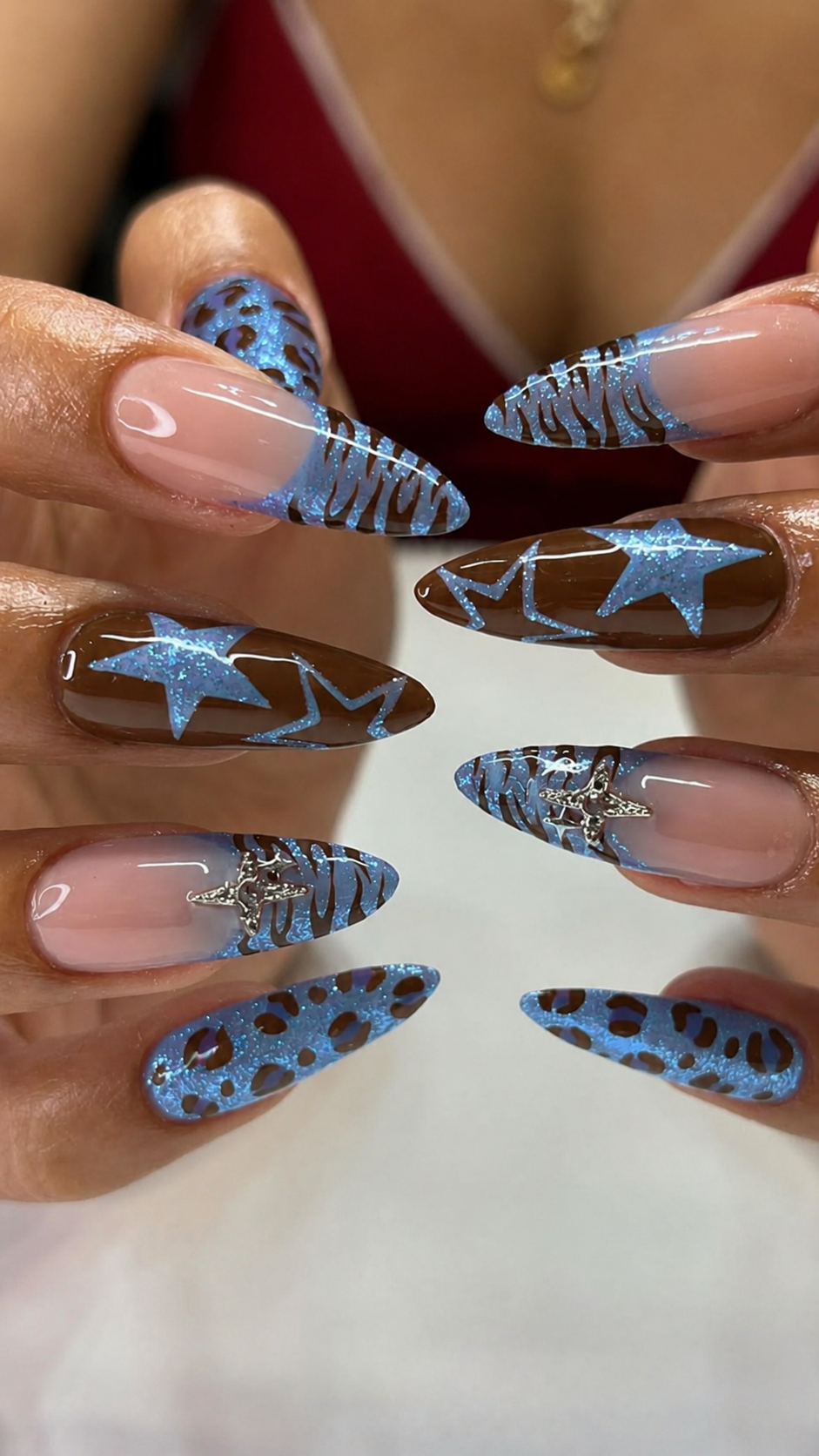 Pointed fingernails display an array of blue and brown designs, including animal prints, glitter stars, and metallic accents.