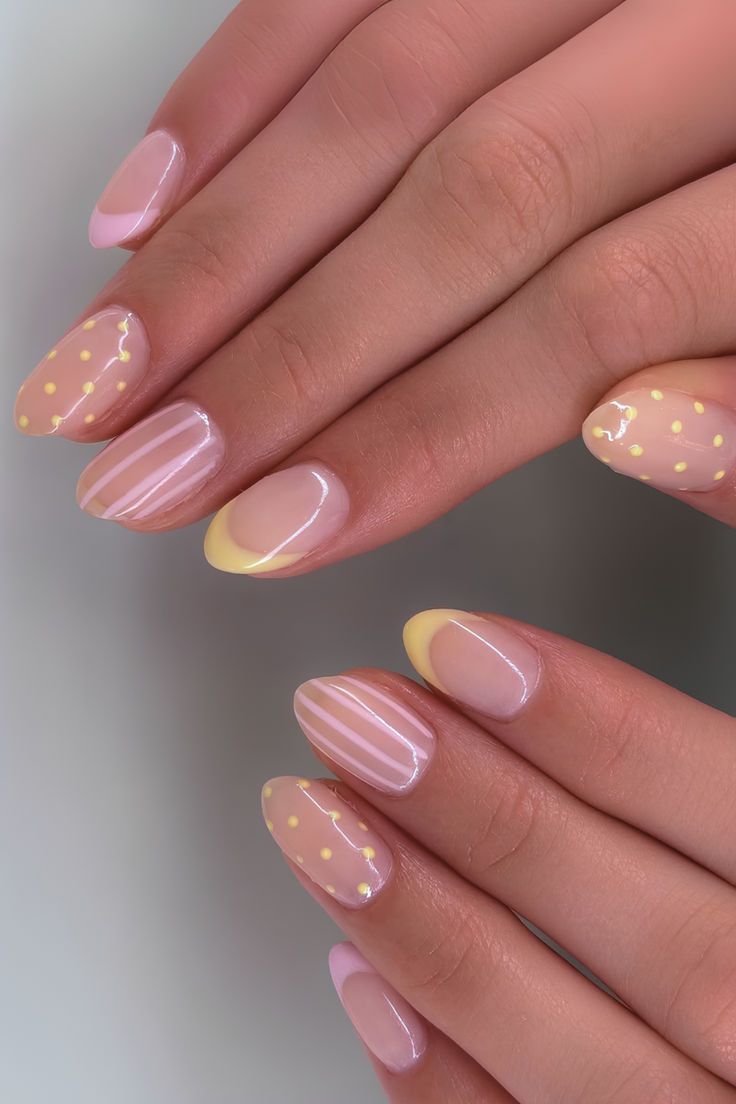 Close-up of manicured hands with short, rounded nails featuring light pink and yellow designs, including polka dots…