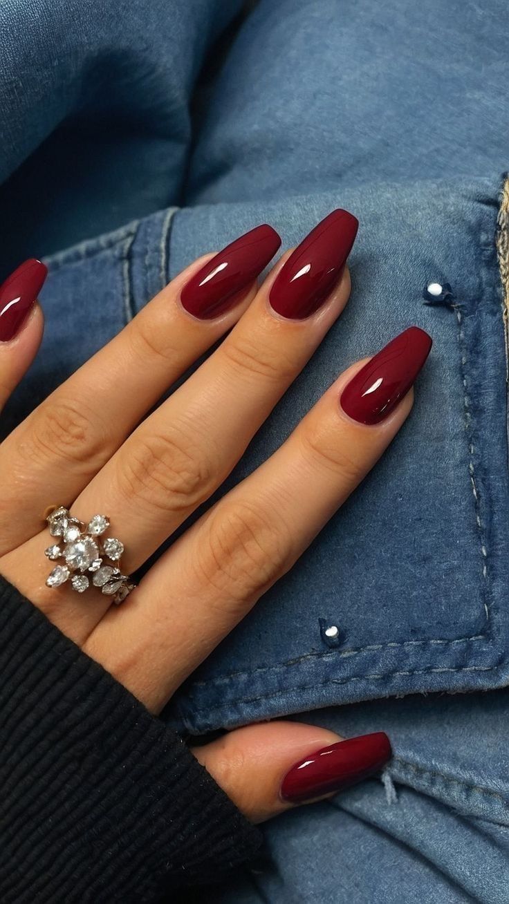 Hand with long, glossy, dark red coffin-shaped nails and a diamond ring against a denim jacket sleeve.