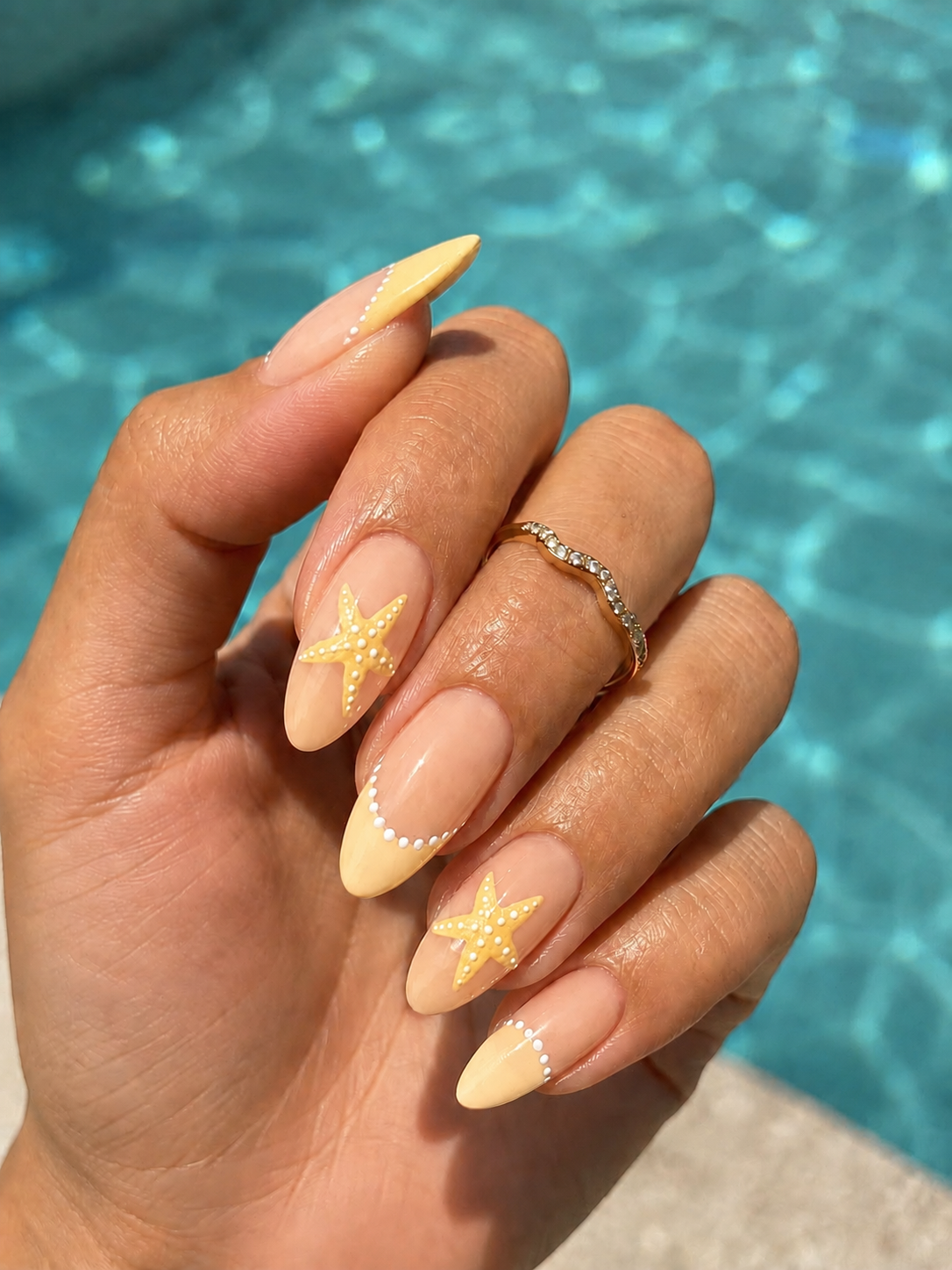 Close-up of a hand with almond-shaped nails featuring yellow French tips, white dots along the tips, and starfish art on two…