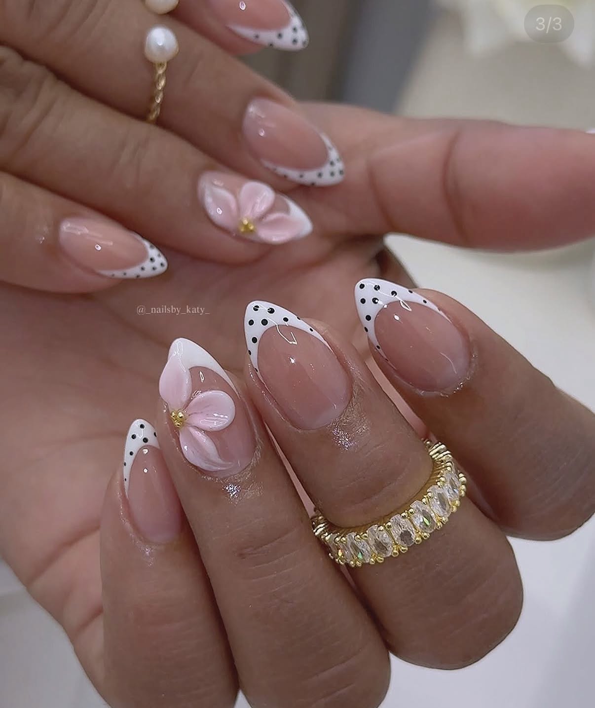 Close up of a hand with almond-shaped nails featuring French tips with polka dots and 3D floral designs.