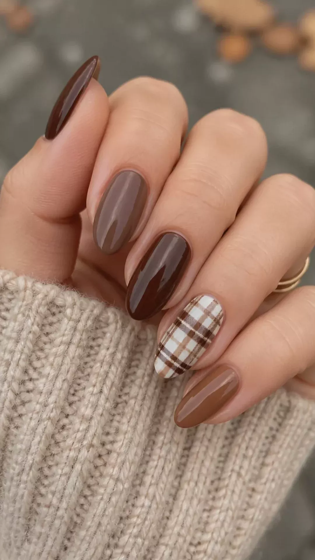 Hand with oval nails, three in brown, one in dark brown, and one with a brown and white plaid design.