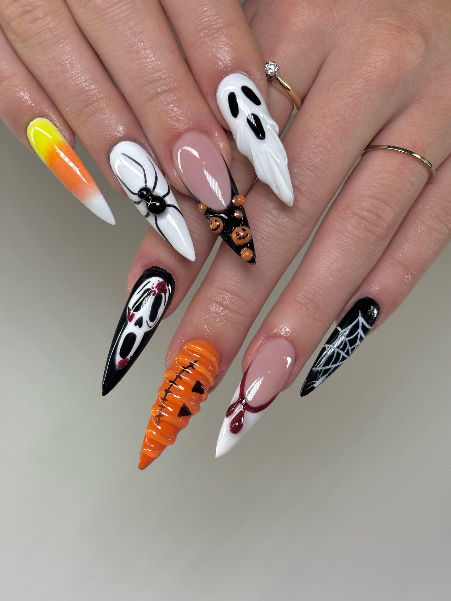 Close-up of a hand with long, pointed stiletto nails, each featuring a different Halloween design including a ghost, spider…