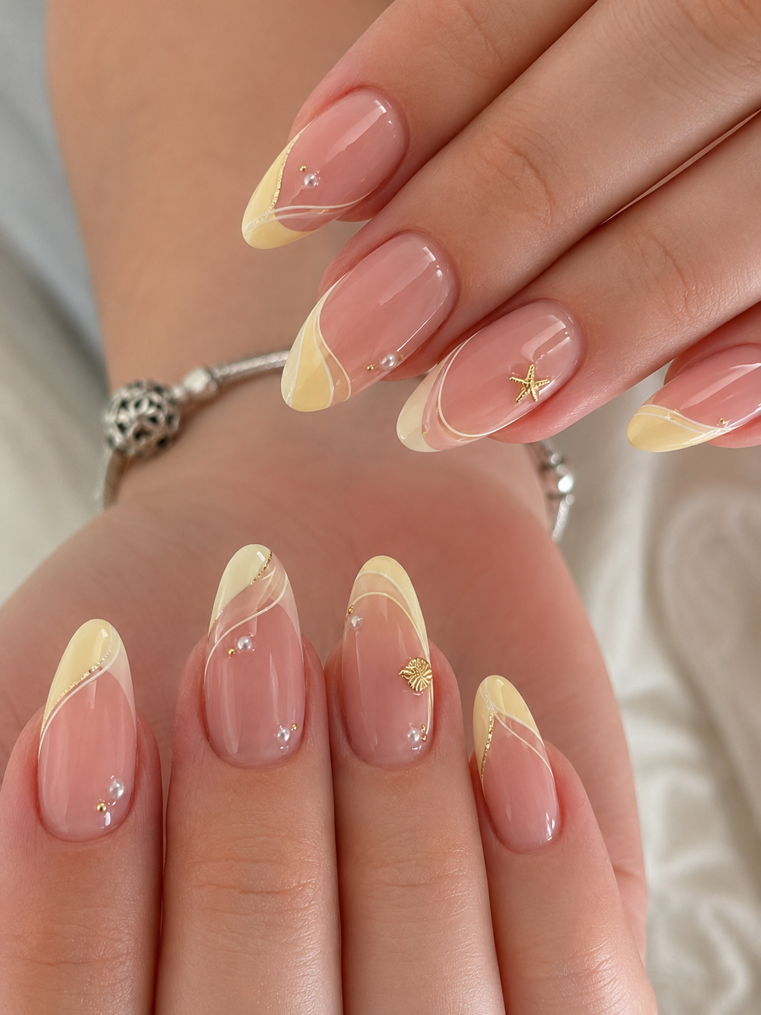 Close up of manicured hands with almond-shaped nails featuring yellow tips, wavy white lines, small pearls, and gold…
